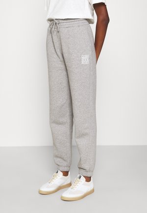 Persona vistiendo pantalones deportivos grises con cintura de cordón y logotipo de texto blanco en el muslo, combinado con zapatillas blancas sobre fondo claro.