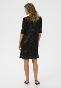 Un vestido negro de lentejuelas con mangas cortas y un ajuste holgado. La tela cuenta con una textura brillante en toda su superficie, terminando a medio muslo.