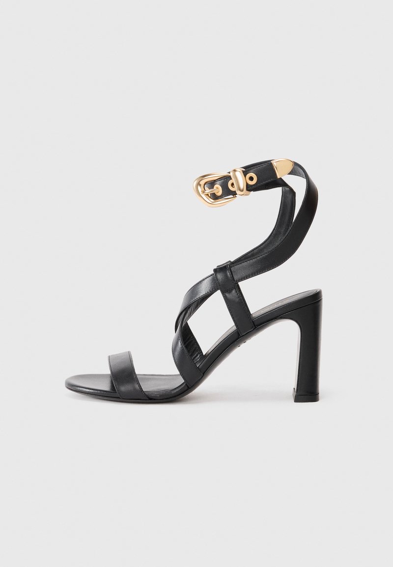 Zwarte leren sandalen met hoge hak met kruislings gespen en een gouden gespsluiting rond de enkel, op een effen lichte achtergrond.