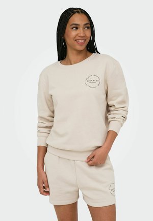 Kvinde med flettet hår iført beige sweatshirt og matchende shorts med trykte logoer, står og smiler med hånden i lommen.
