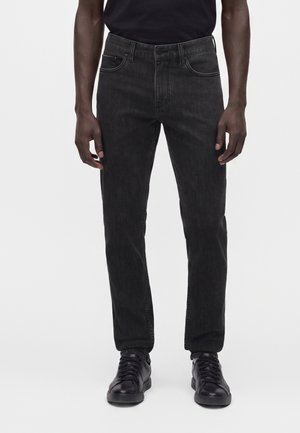 Mann trägt schwarze Slim-Fit-Jeans und schwarze Sneakers, steht vor einem schlichten weißen Hintergrund.