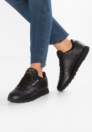 Person iført sorte Reebok Classic sneakers og blå jeans med flosset kant, stående på en hvid overflade.