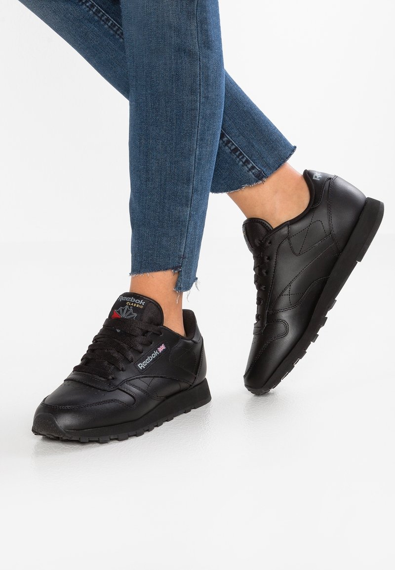 Personne portant des baskets Reebok Classic noires et un jean bleu avec des ourlets effilochés, debout sur une surface blanche.