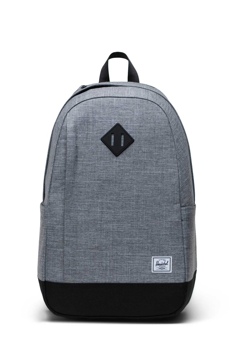 Mochila de tela gris con una textura suave, forma redondeada, fondo negro y un parche en forma de diamante en la parte frontal que presenta dos líneas verticales.