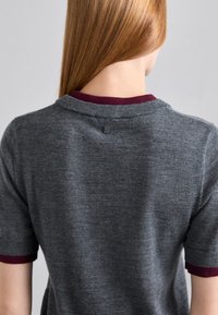Personne aux longs cheveux roux portant un pull gris à manches courtes avec un bord bordeaux au niveau du col et des manches, vue de dos.
