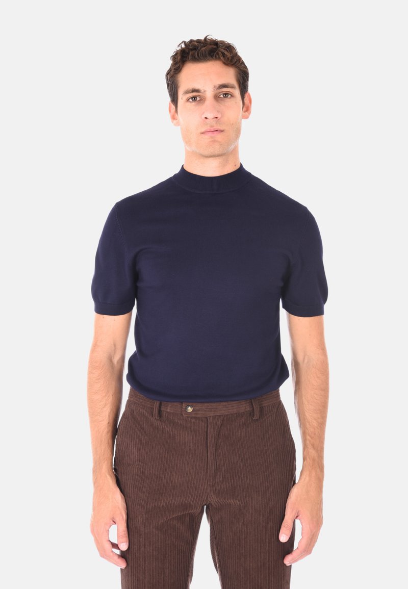 Uomo che indossa un maglione a collo alto corto blu navy e pantaloni in corduroy marroni, in piedi contro uno sfondo semplice.