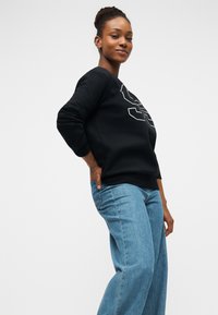 Μαύρο φούτερ με υπερμεγέθη γραφικό, στρογγυλή λαιμόκοψη και ριμπ μανσέτες. Συνδυασμένο με ανοιχτό γαλάζιο τζιν σε φαρδιά γραμμή. Λείο υφής ύφασμα.