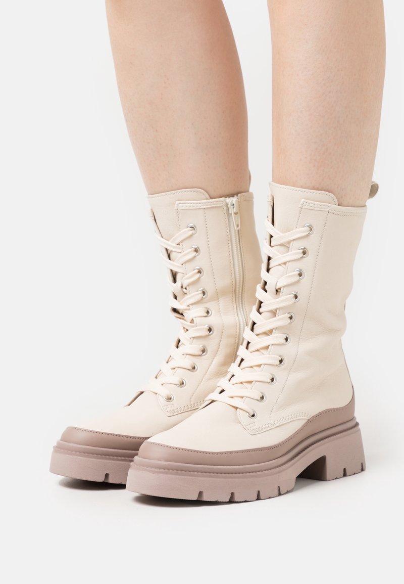Bottes mi-mollet crème à lacets avec fermetures éclair latérales et semelles plateformes épaisses couleur taupe, portées sur jambes nues.