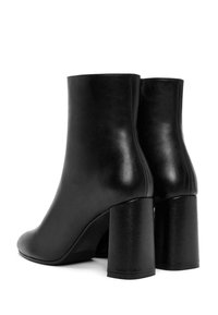 Botas de tobillo de cuero negro con textura suave, punta redondeada y tacón alto grueso. La parte trasera presenta costuras sin juntas.
