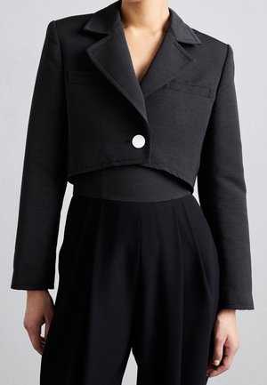 Mujer con una blazer negra corta texturizada con un botón blanco y pantalones negros de talle alto.