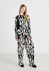 Une tenue plissée noir et blanc présente des motifs abstraits ; manches longues, pantalons larges et un sac à bandoulière texturé. Des chaussures noires complètent le look.