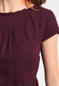 Robe bordeaux à manches courtes, présentant des plis au niveau de l'encolure et un tissu lisse et texturé. Design simple et élégant avec un corsage ajusté.
