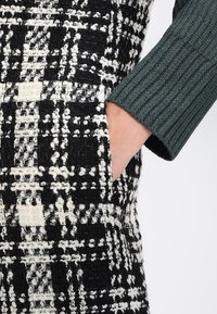 Tkana tkanina z czarno-białym wzorem w kratę, o miękkiej, teksturowanej powierzchni. Częściowo widoczny zielony rękaw z prążkowaną fakturą.