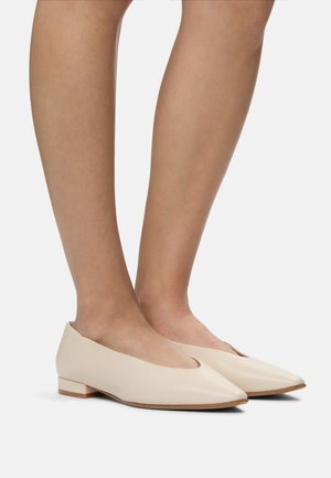Beige mokassins dari kulit dengan ujung runcing, tumit blok rendah, dan sisi yang dipotong, menampilkan tekstur halus dan desain minimalis.