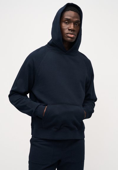 Tmavě modrá mikina s kapucí a volnějším střihem, s žebrovanými manžetami, předními kapsami a měkkou texturou. Design má raglánové rukávy.