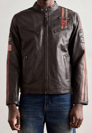 Man die een donkerbruine leren jas draagt met rode en beige racestrepen en meerdere motorfietsthema-patches, gecombineerd met een blauwe spijkerbroek.