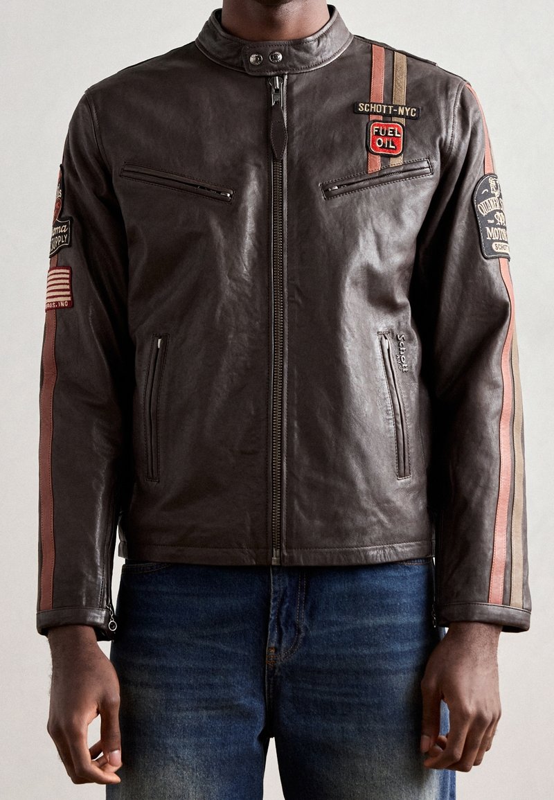 Homme portant une veste en cuir marron foncé avec des bandes de course rouges et beiges et plusieurs patchs à thème moto, associé à un jean bleu.