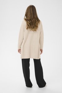 Beige cable-knit neulepaita pitkillä hihoilla, jossa on teksturoitu kuvio ja löysä malli, yhdistettynä mustiin leveälahkeisiin housuihin.