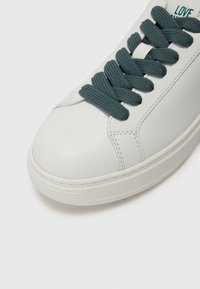 Balti ādas čības ar noapaļotu purngalu, kurām ir pelēki auduma šņores, izšūtas detaļas un teksturēta gumijas zole.