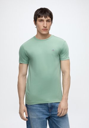 Jeune homme aux cheveux courts et foncés portant un t-shirt moulant vert clair et un jean bleu, debout devant un fond blanc uni.