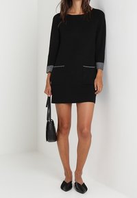 Abito in maglia nera con maniche a tre quarti, rifinitura grigia sulle tasche e due tasche frontali. Abbinato a scarpe ballet flat nere e una borsa a tracolla piccola.