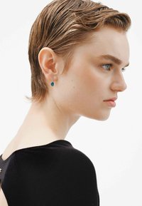 Pendientes de gota con una gema azul, rodeada de pequeños cristales transparentes, que cuelgan de un delicado engaste de plata. Cuello de prenda negra visible.