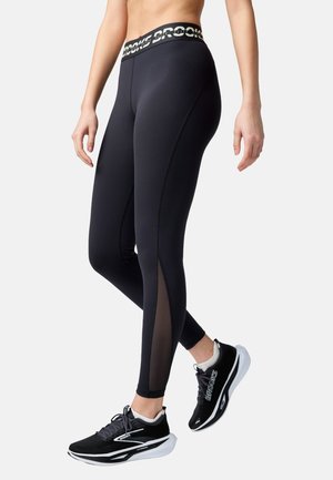 Female wearing black high-waist leggings with mesh side panels and black and white Brooks running shoes, standing with legs crossed.