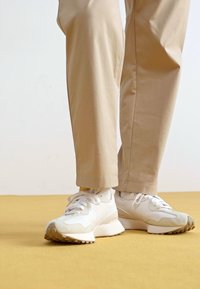 Weiße sportliche Sneaker mit strukturierten Akzenten, Schnürdesign und robusten Gummisohlen, kombiniert mit hellbeigen Hosen vor einem gelben Hintergrund.