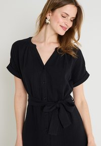 Vestido negro de manga corta con escote en v, botones delanteros y cinturón anudado en la cintura. Hecho de una tela texturizada. Presenta mangas enrolladas.