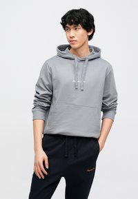 Hall hallitud kapuutsiga dressipluus, millel on reguleeritavad nööbid, kängurutasku ja soonikkoes varrukaotsad, sobitatud tumeliste jõogapükstega, millel on oranž tikand.