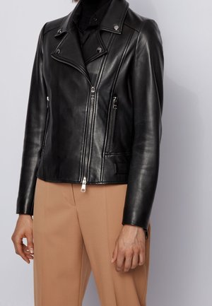 Zwarte leren bikerjack met ritssluitingen en drukknopen, gedragen met beige broek bij een persoon.