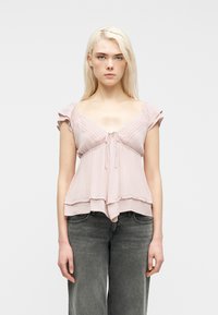 Νεαρή γυναίκα με μακριά ξανθά μαλλιά, φορώντας ένα ανοιχτό ροζ πουκάμισο με βολάν και ξεβαμμένο μαύρο τζιν, στέκεται μπροστά σε μια απλή λευκή επιφάνεια.