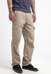 Beige cargo pants with multiple side pockets, made from cotton fabric. Relaxed fit with a slightly wrinkled texture and reinforced seams.