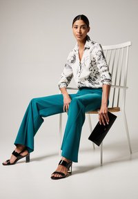 Witte blouse met zwart bloemenprint, teal wijduitlopende broek, zwarte hoge sandalen en een zwarte clutch. Model zit op een witte stoel.