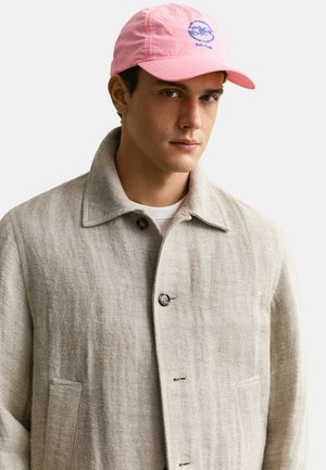 Jonge man met een beige gestructureerd jasje met knopen en een roze baseballpet met blauwe borduursels op een lichte achtergrond.