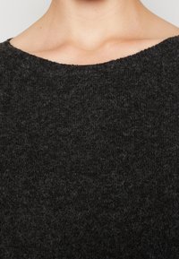 Detailní záběr horní části hrudníku osoby v texturovaném, tmavě šedém pleteném svetru s širokým výstřihem, na světlem pozadí.