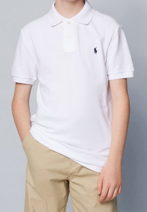 Barn iført en hvid kortærmet poloshirt med en lille mørk logo og beige bukser, stående foran en ensfarvet baggrund.
