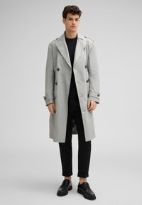 Doppelreihiger Trenchcoat aus hellgrauem Stoff mit Kragen, großen Knöpfen und Schulterklappen, kombiniert mit schwarzen Hosen und Schuhen.