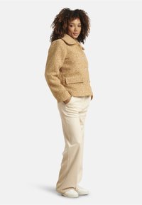 Fitted tan wool-blend jacket with a collar, front buttons, and side pockets, paired with cream wide-leg trousers and white sneakers.