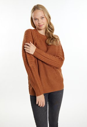 Dicker Strickpullover in warmem Braun mit strukturiertem Muster, gerippten Bündchen und seitlichen Schlitzen. Kombiniert mit dunklen, enganliegenden Jeans.