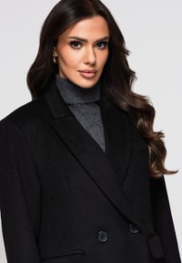 Svart dubbelknäppt kappa av texturerat tyg med slag i kragen och två framfickor. Bärs över en mörkgrå polotröja.