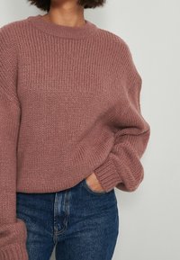 Jasnoróżowy, gruby sweter w splot o okrągłym dekolcie i luźnym kroju. Prążkowana faktura i szerokie rękawy, zestawiony z niebieskimi dżinsami.