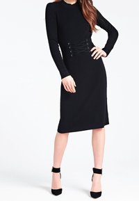 Femme portant une robe midi côtelée noire à manches longues avec un détail lacé à la taille et des escarpins noirs à bride à la cheville, debout avec une main sur la hanche.