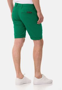 Grüne Shorts im Slim-Fit-Stil, mit hochgeschlagenen Säumen, Gesäßtaschen und roten Steppnähten. Kombiniert mit weißen Sneakers.