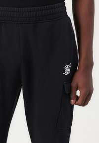 Black sweatpants made of soft fabric, featuring an elastic waistband, side cargo pocket, and white embroidered logo on the thigh.