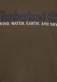 Donkergroen olijfgroen katoenen t-shirt met een gedurfde marineblauwe "Timberland"-logo en witte tekst met de woorden "WIND, WATER, EARTH, AND SKY." Gladde textuur.