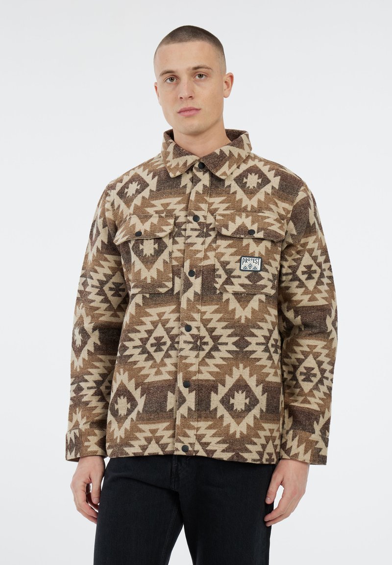 Man in een bruine en beige geometrisch patroon jack met knopen, voorzien van twee voorzakken en een klein "Protest" logo patch.