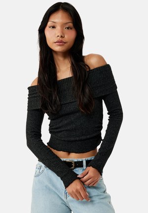 Femme aux longs cheveux foncés portant un pull côtelé noir à épaules dénudées et un jean bleu clair avec une ceinture noire, debout devant un fond blanc.