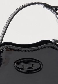 Čierna kabelka z lakovanej kože s vlnitými okrajmi, oválnym zapínaním a krátkymi dvojitými rúčkami na jednoduchom pozadí.