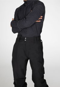 Personne portant un pantalon noir taille haute avec des boutons argentés et un haut sombre à manches longues zippé, bras croisés devant un fond uni.